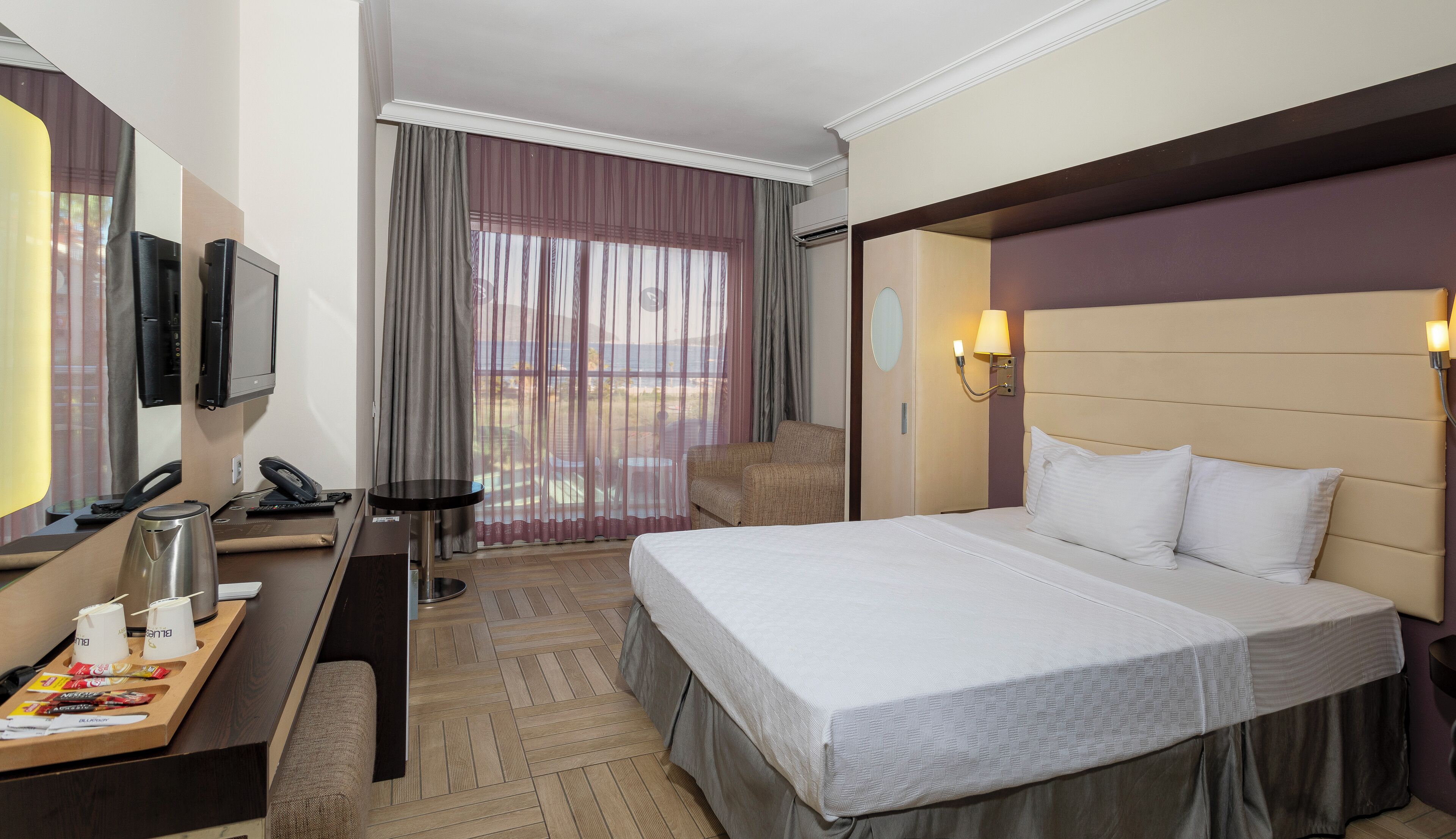 Habitació d'hotel amb un llit, una escriptori amb telèfon i un fabricant de cafè, una cadira i una finestra amb cortines.