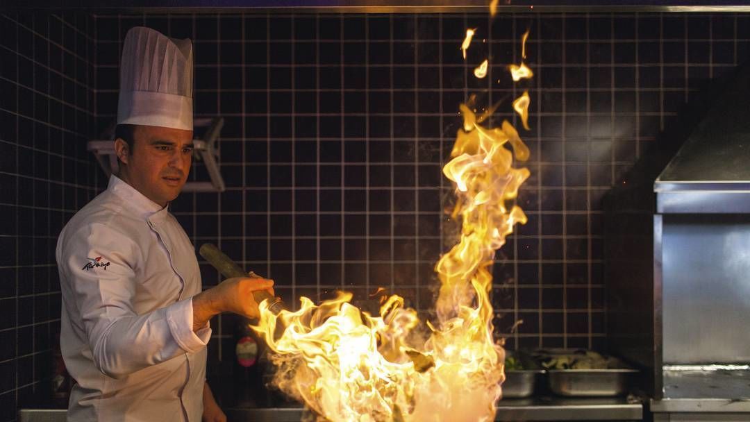 A chef performing a flambé with flames rising from a pan.