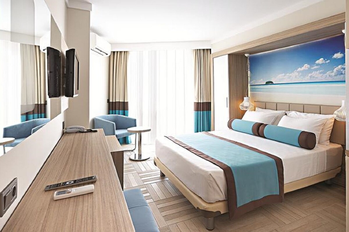 A hotel room with a bed, a desk, a chair, a TV, and a window with a beach view.
