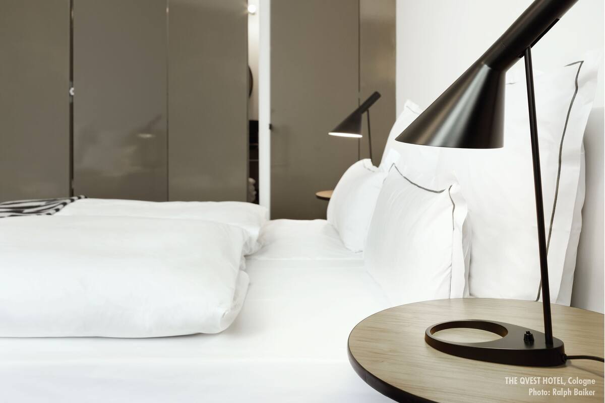 Ein modernes Hotelzimmer mit einem ordentlich bezogenen Bett, einem Nachttisch mit Lampe und Blick auf einen Kleiderschrank.