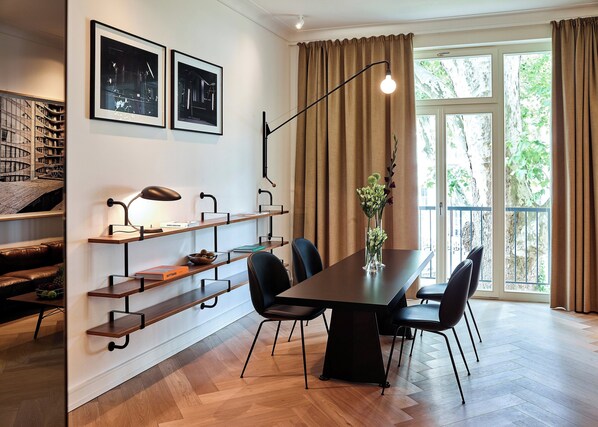 Un comedor moderno con una mesa de madera, sillas negras y un jarrón con flores. En la pared, hay estantes con libros y una lámpara.