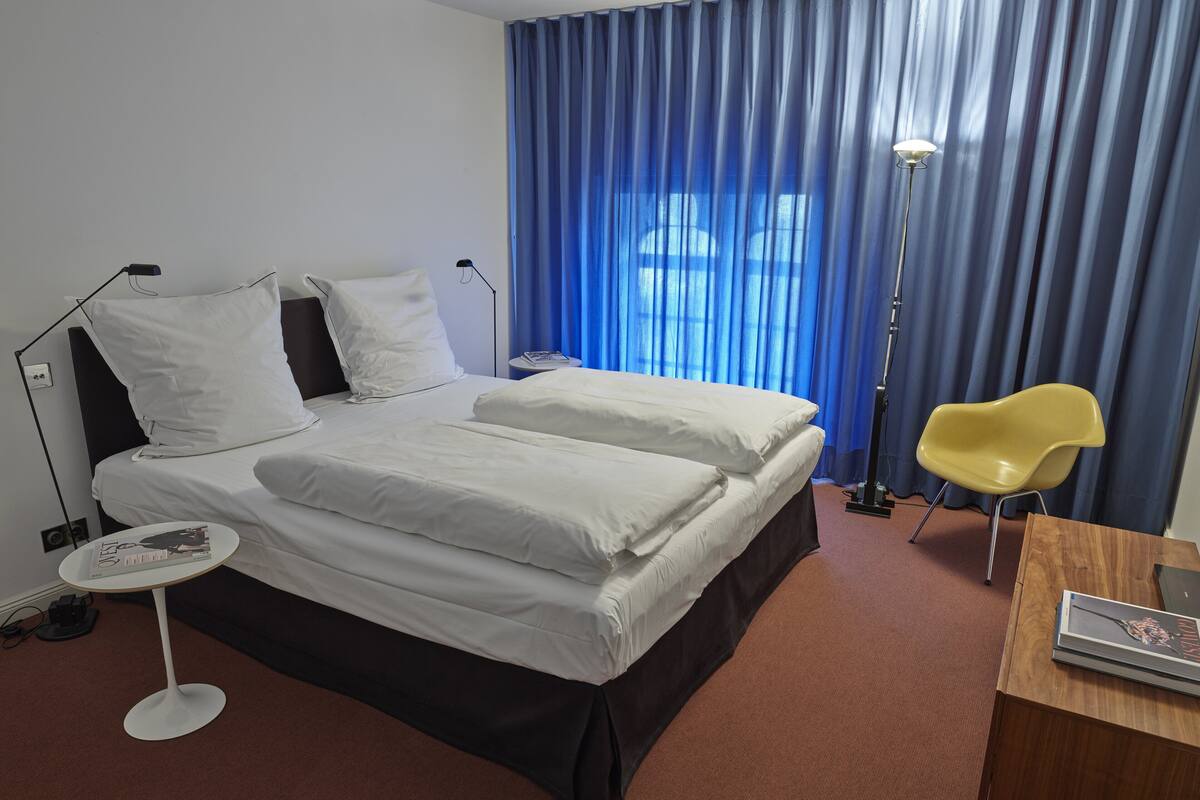 Ein Hotelzimmer mit einem Bett, einem Nachttisch mit einer Zeitschrift, einer Lampe, einem Sessel und einem Fenster mit blauen Vorhängen.