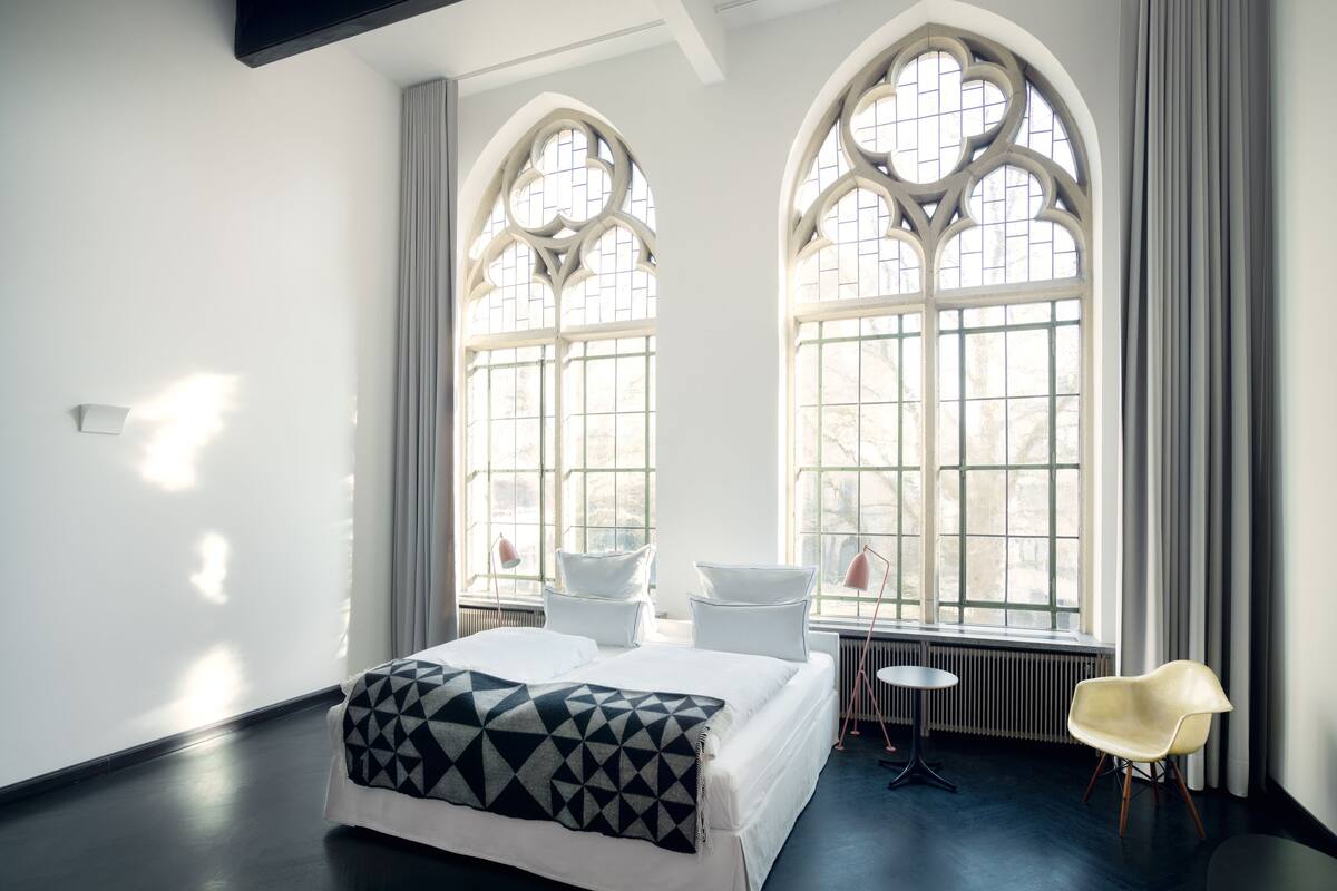 Ein modernes Schlafzimmer mit einem großen Bett, zwei Fenstern mit aufwendigen Verzierungen, einem kleinen Tisch und einem Stuhl.