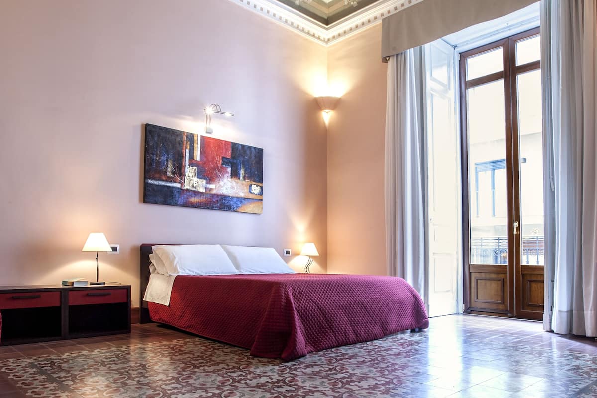 A hotel room with a large bed, two bedside tables with lamps, a patterned rug, and a balcony with curtains.