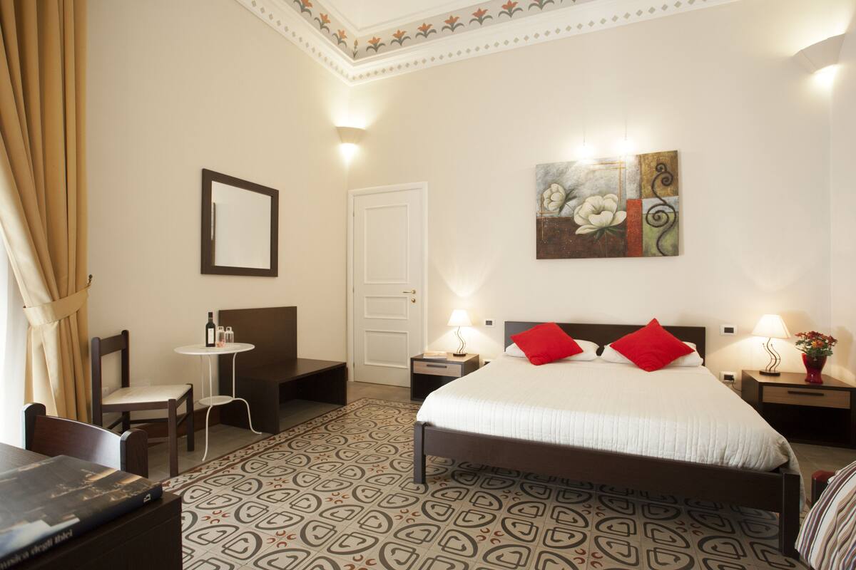 A hotel room with a bed, bedside tables, a chair, a desk, a mirror, and a painting on the wall.
