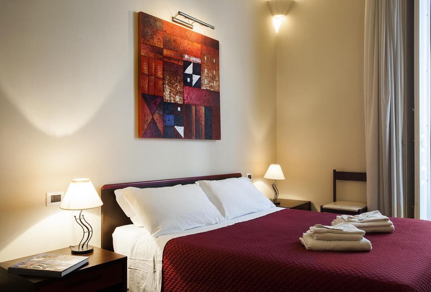 Quarto de hotel com uma cama grande, duas luminárias de cabeceira, uma obra de arte na parede e vista para o exterior através de uma janela.