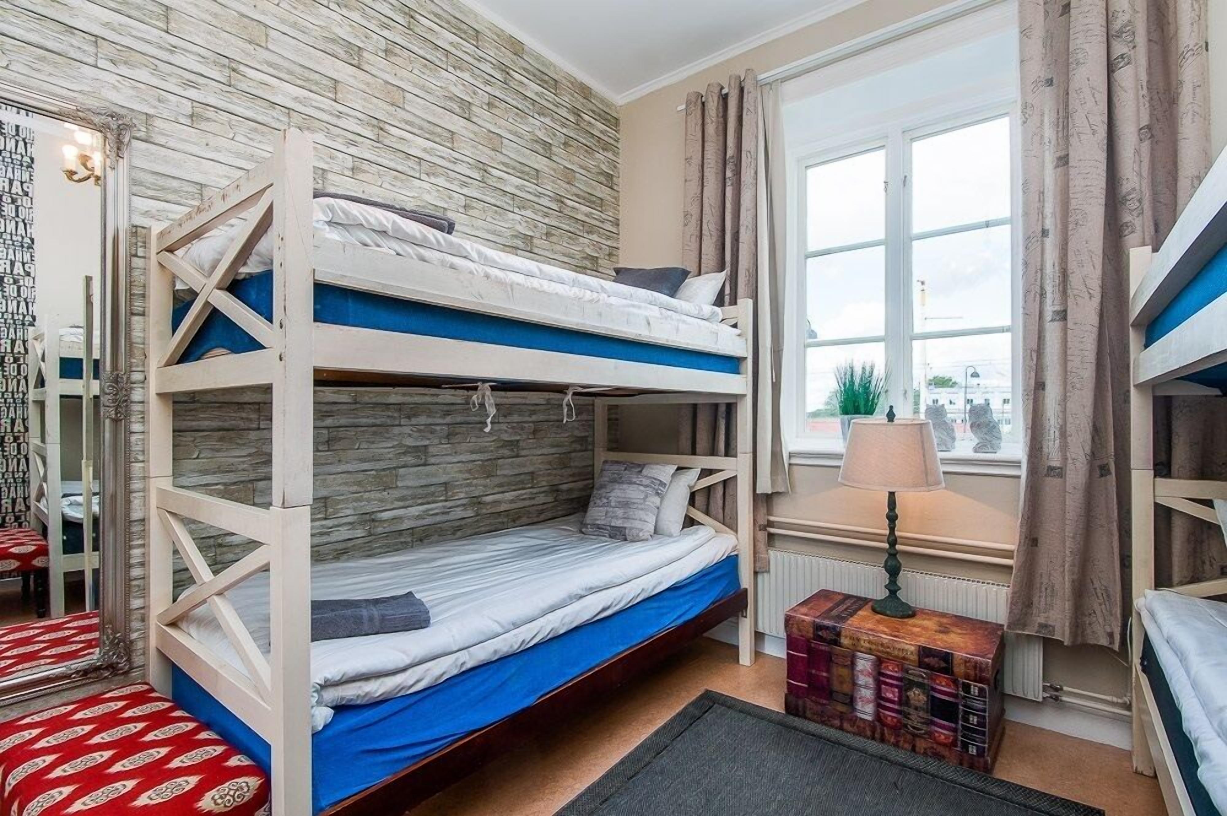 A room with three bunk beds, a window with curtains, a radiator, and a lamp.