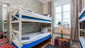 A room with three bunk beds, a window with curtains, a radiator, and a lamp.