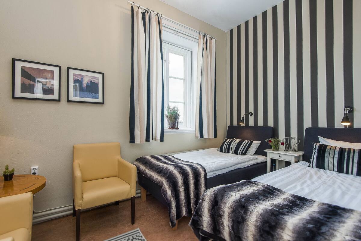 Ein Schlafzimmer mit zwei Betten, einem Sessel, einem kleinen Tisch und einem Fenster mit Vorhängen.