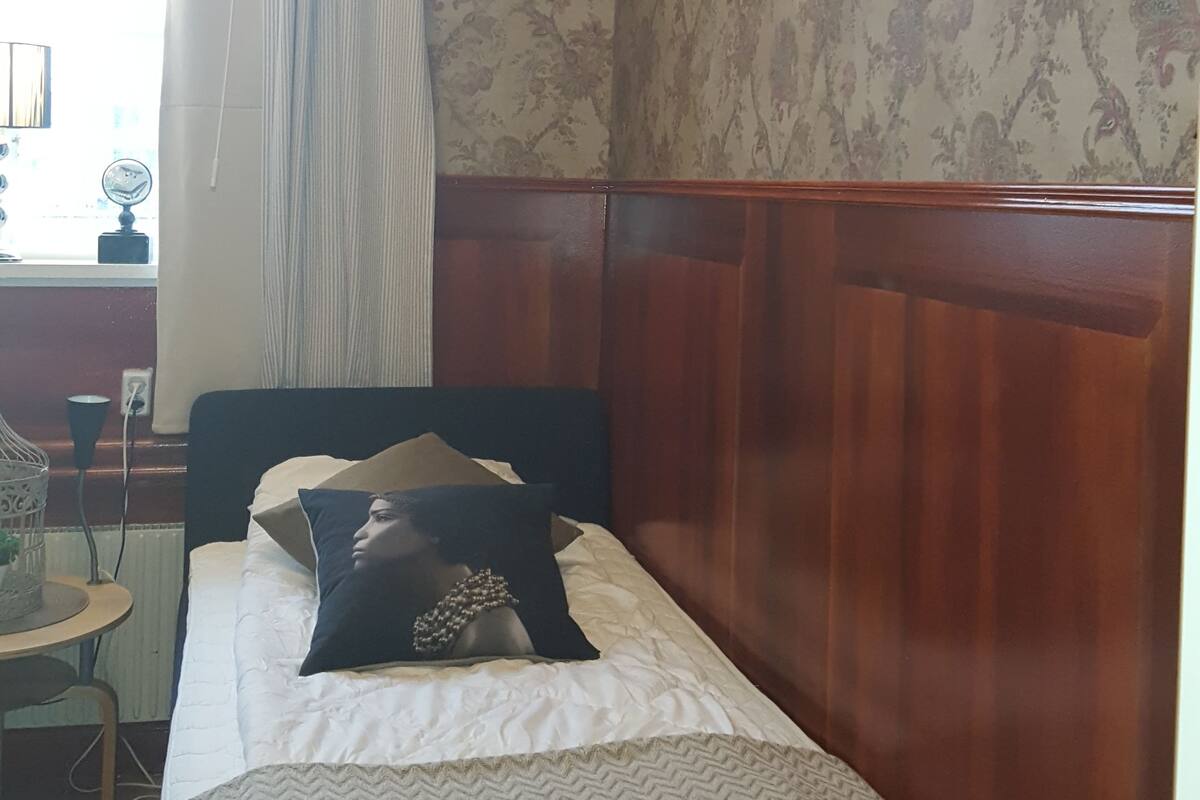Ein Schlafzimmer mit einem Holzkopfteil, einem Bett mit einem gemusterten Bettbezug, einem Fenster mit Vorhängen und einer Wand mit floralem Tapetenmuster.