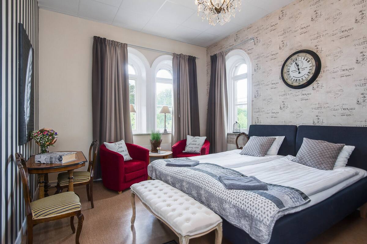 Ein Hotelzimmer mit einem großen Bett, zwei roten Sesseln, einem kleinen Tisch mit Blumenvase und einer Uhr an der Wand.