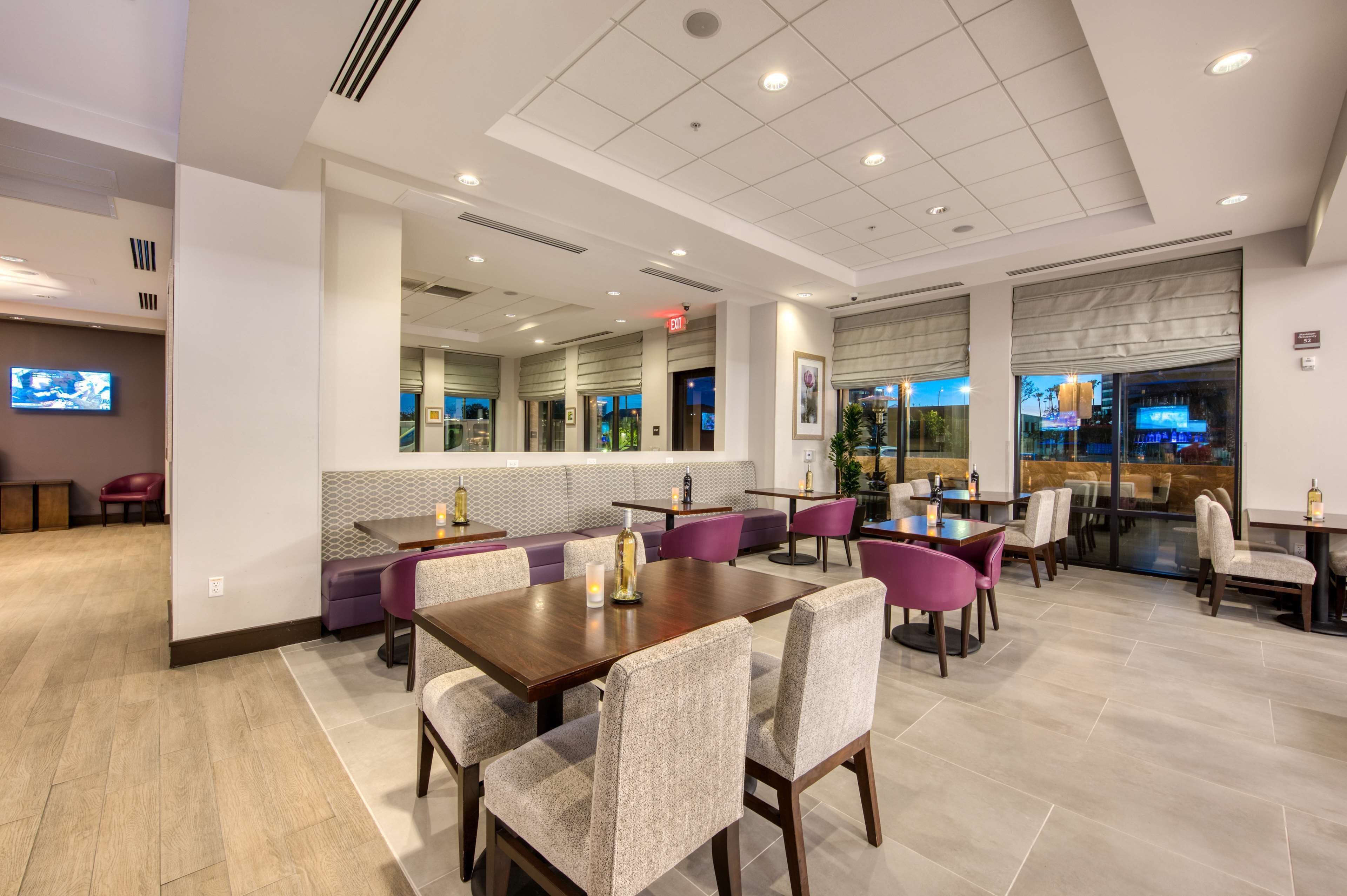 A modern dining area with multiple tables and chairs, a seating area with cushioned benches, and large windows providing views of the outside.
