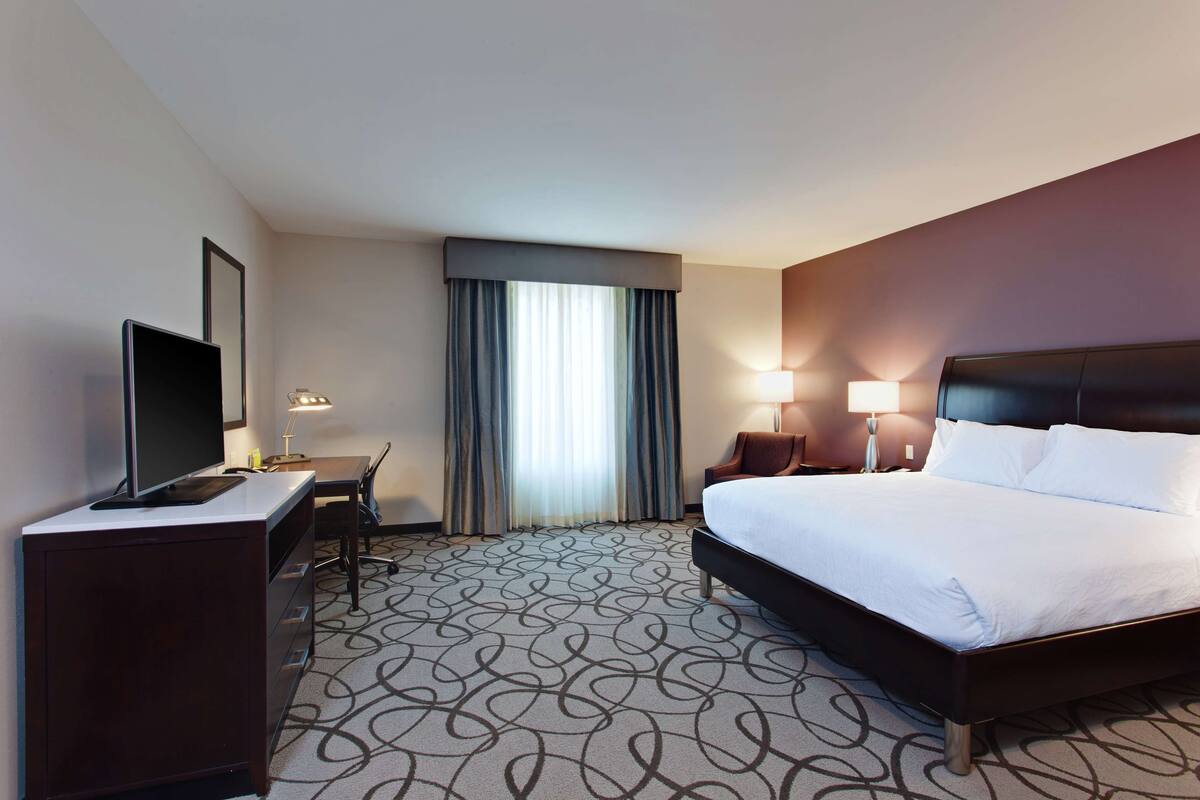 Ein Hotelzimmer mit Bett, Schreibtisch mit Computer, Fernseher und einem Fenster mit Vorhängen.