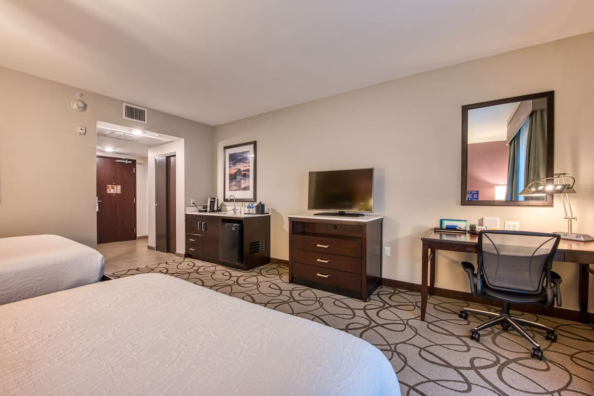 Ein Hotelzimmer mit zwei Betten, einem Schreibtisch mit Stuhl, einem Fernseher, einer Minibar und einer Badezimmertür.