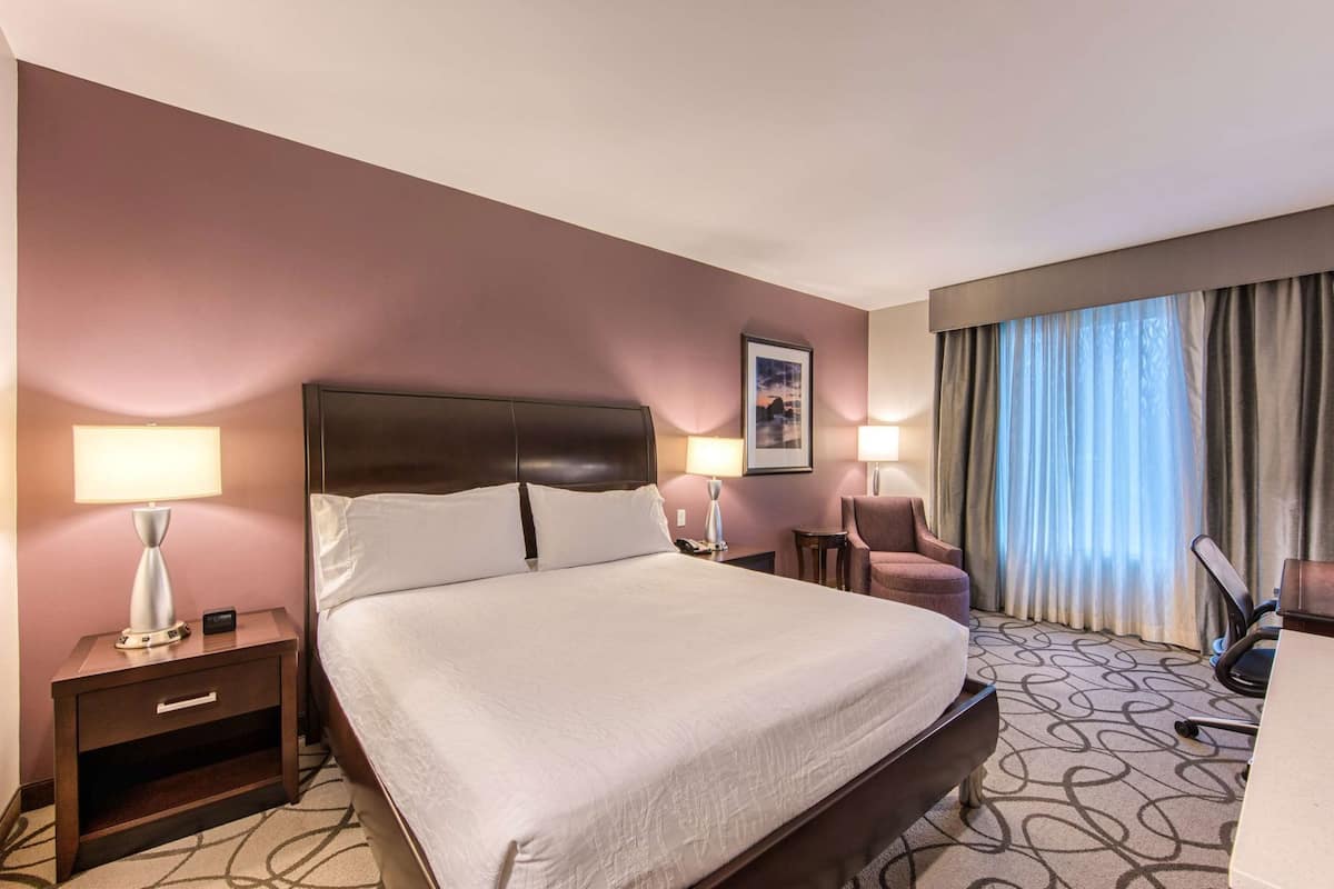 Ein Hotelzimmer mit einem großen Bett, Nachttischen mit Lampen, einem Sessel, einem Schreibtisch und einem Fenster mit Vorhängen.