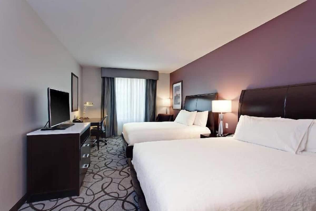Ein Hotelzimmer mit zwei Betten, einem Schreibtisch mit Fernseher und einem Fenster mit Vorhängen.