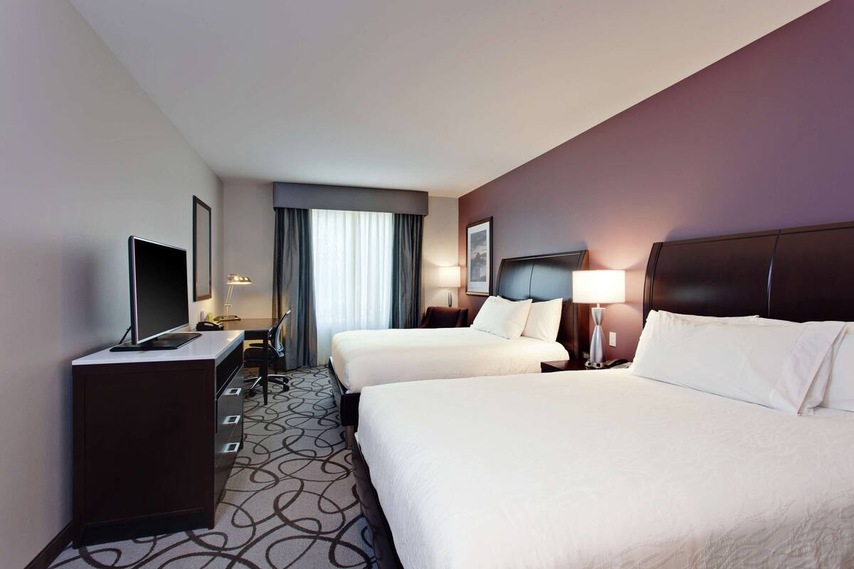Ein Hotelzimmer mit zwei Betten, einem Schreibtisch mit Fernseher, einer Lampe und einem Fenster mit Vorhängen.