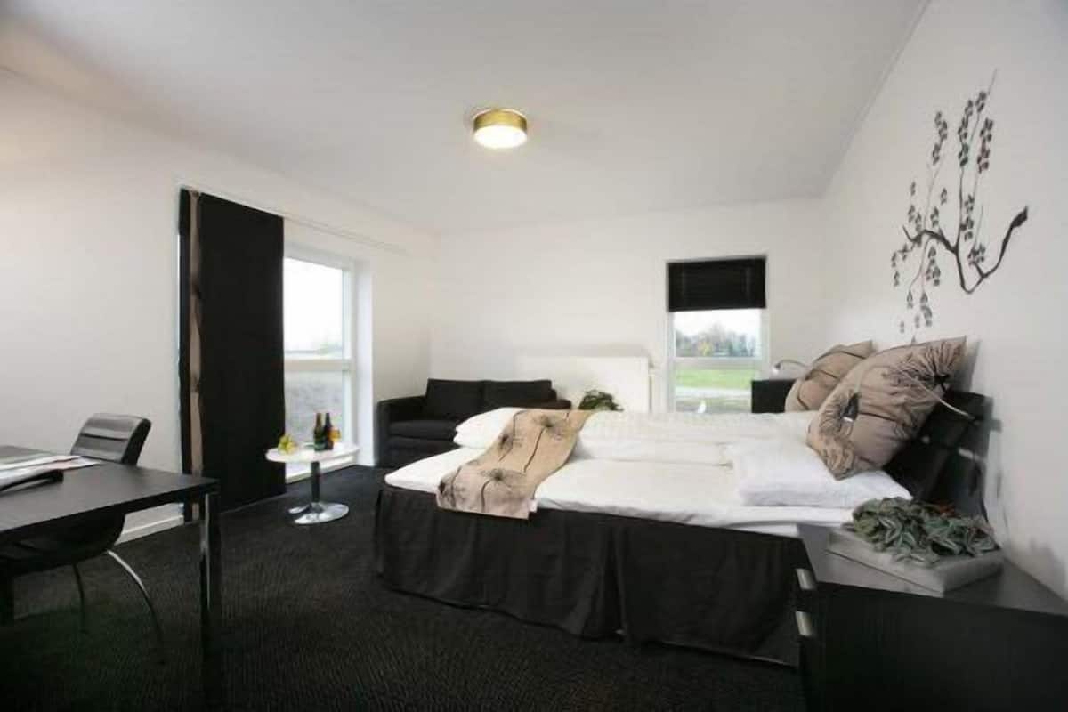 Une chambre d’hôtel moderne, équipée d’un lit, d’un bureau et d’une chaise. Une fenêtre laisse entrer la lumière naturelle et offre une vue sur l’extérieur.