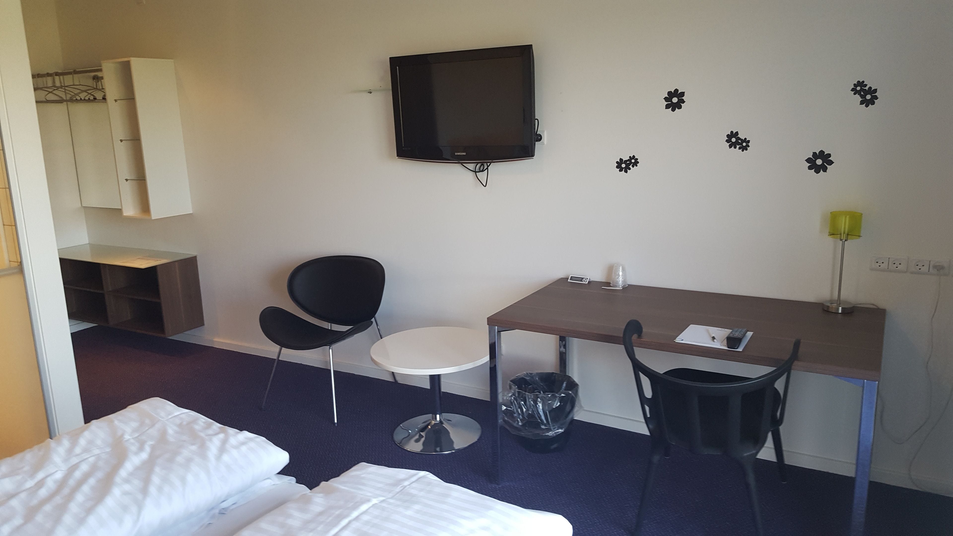 A hotel room with a bed, a desk, a chair, a TV, and a small table.