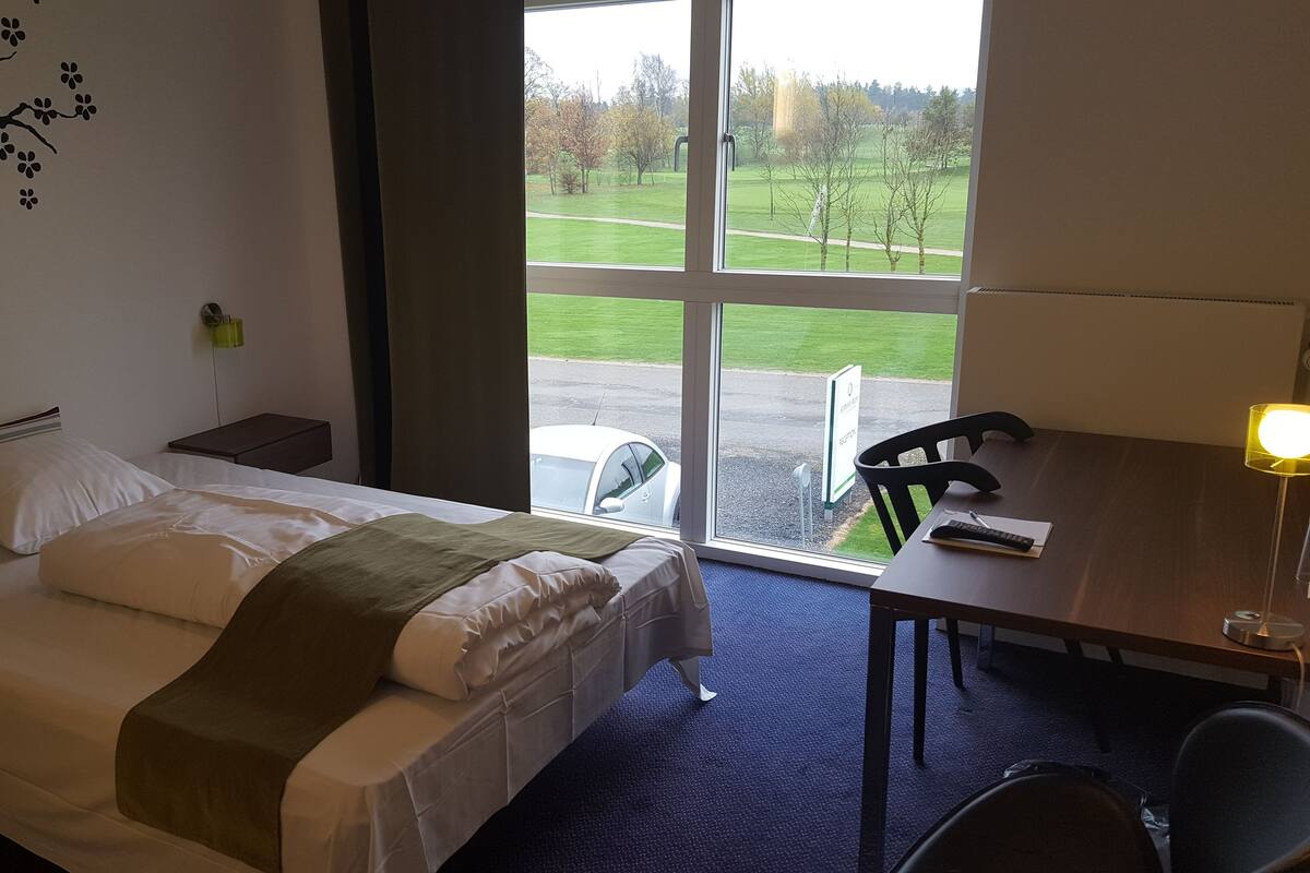 Une chambre d’hôtel équipée d’un lit, d’un bureau, d’une chaise, d’une lampe et offrant une vue sur un parc verdoyant grâce à une grande fenêtre.