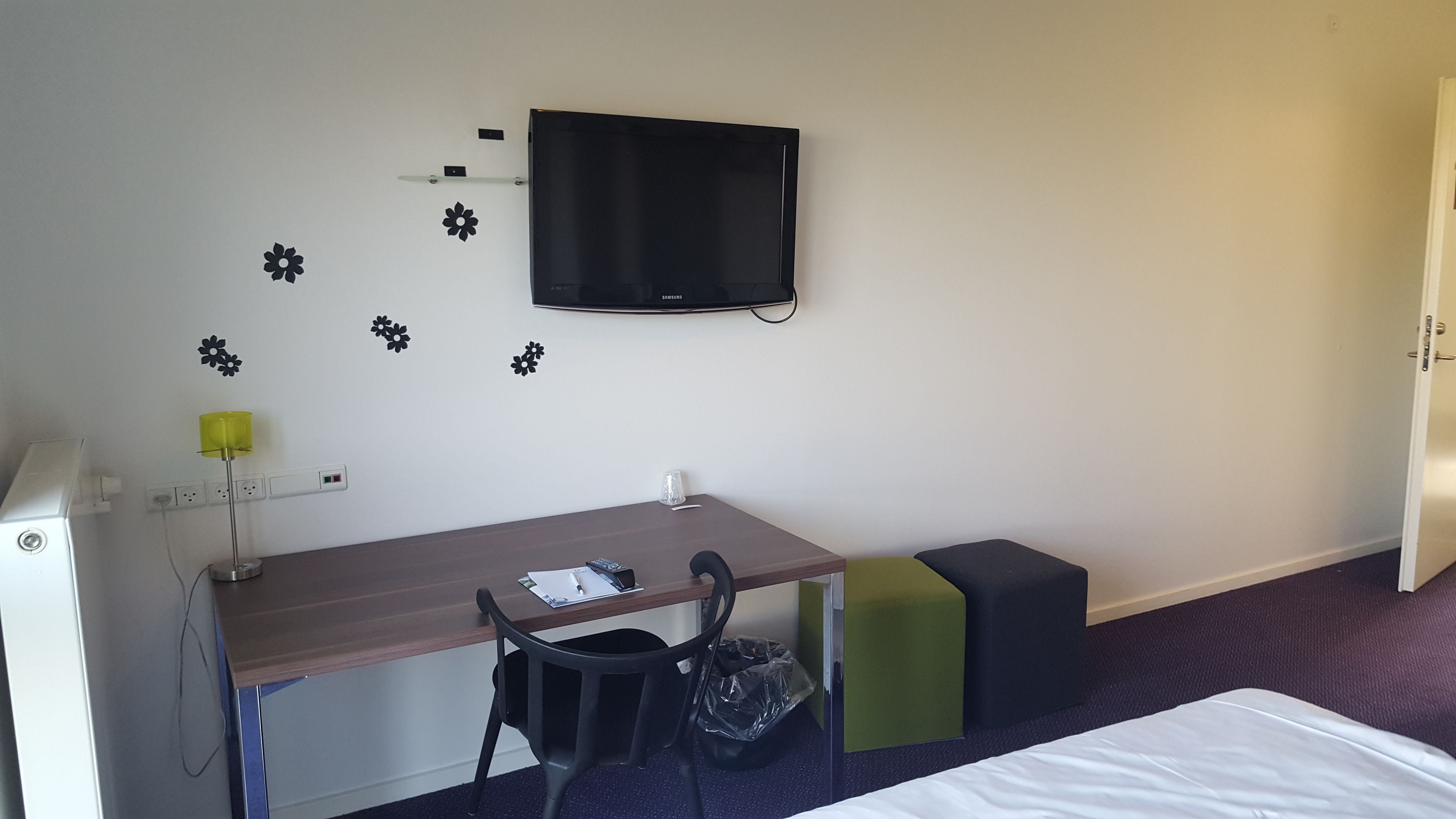 A hotel room with a wooden desk, a chair, a television mounted on the wall, a small shelf, and a bed with a white sheet.