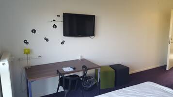 Habitación de hotel con un escritorio de madera, una silla, televisor montado en la pared, un pequeño estante y una cama con sábana blanca.