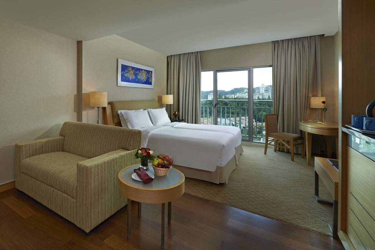 A hotel room with a sofa, a bed, a TV, a small table with a basket of fruit, and a balcony with a view of buildings.