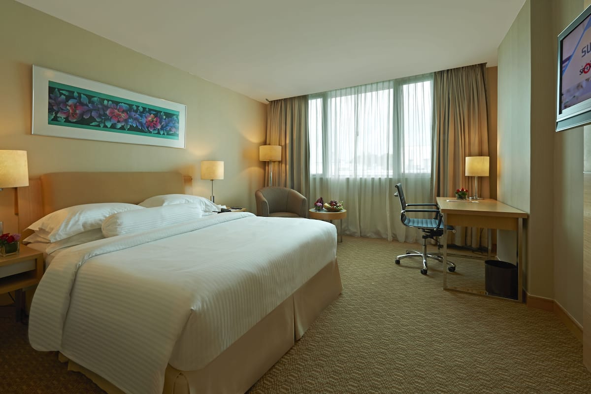 A hotel room with a large bed, two bedside tables with lamps, a desk with a chair, a small side table with a vase, and a large window with curtains.