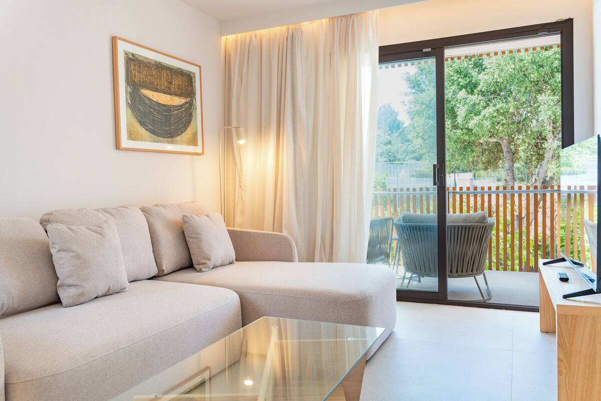 Una sala de estar moderna con un sofá, una mesa de centro de vidrio, un cuadro en la pared y un balcón con vistas a zonas verdes.