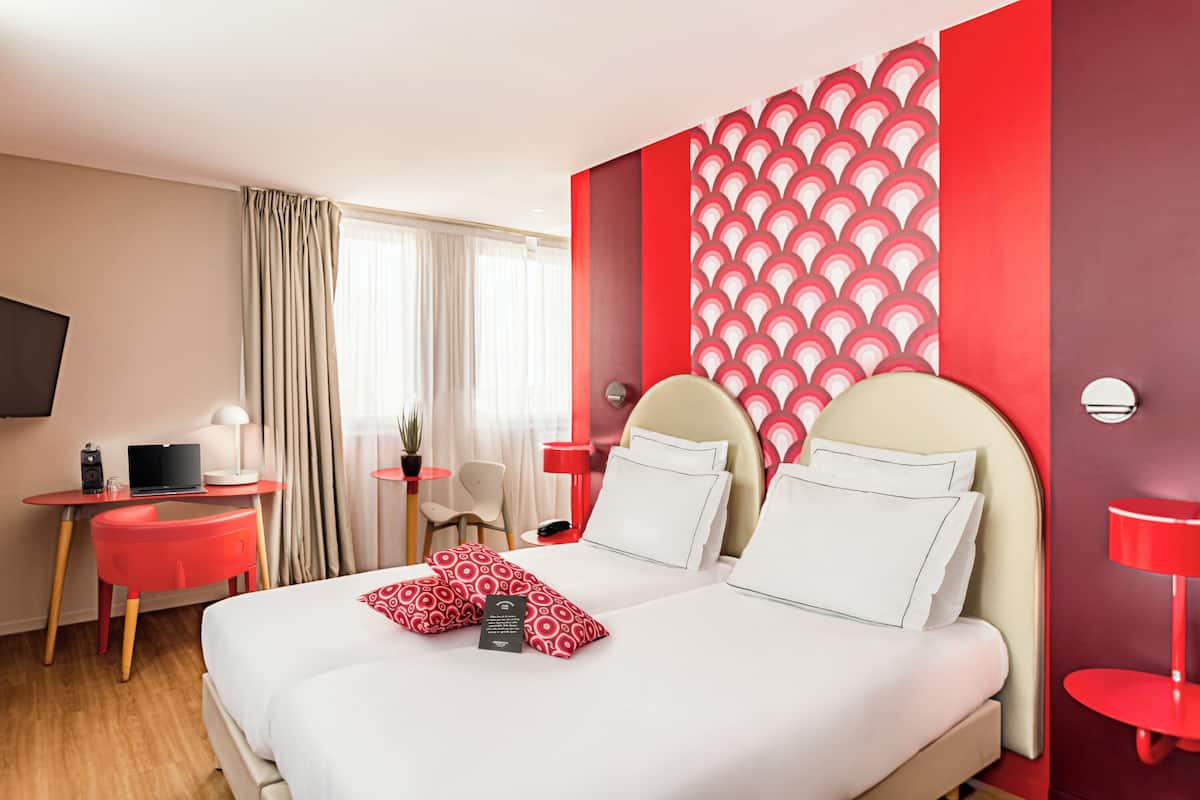 A hotel room with a bed, a desk with a laptop, a chair, a TV, and a red accent wall.