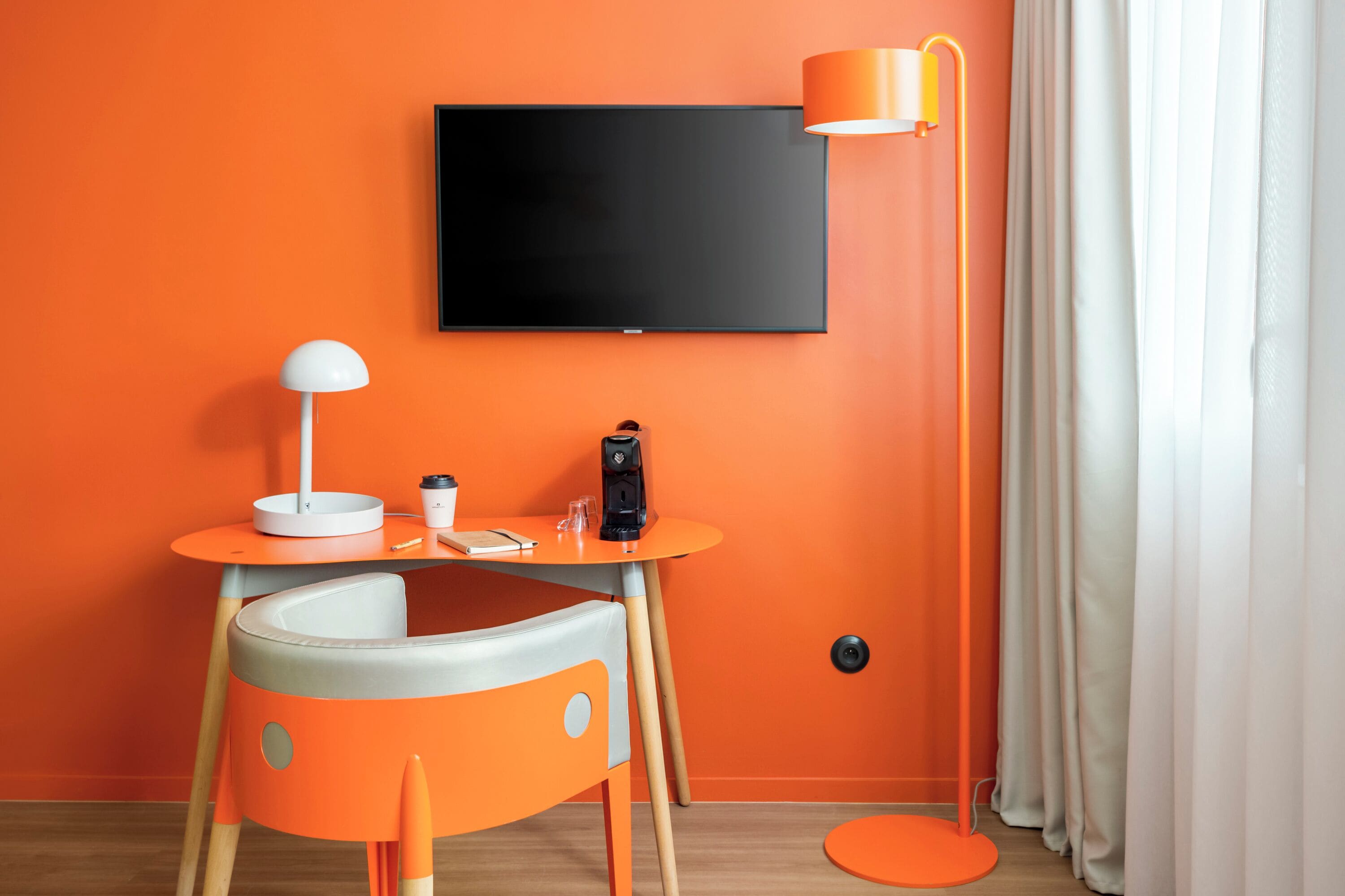 A room with an orange wall, a flat-screen TV mounted above a small table with a lamp, a chair, and a coffee maker.