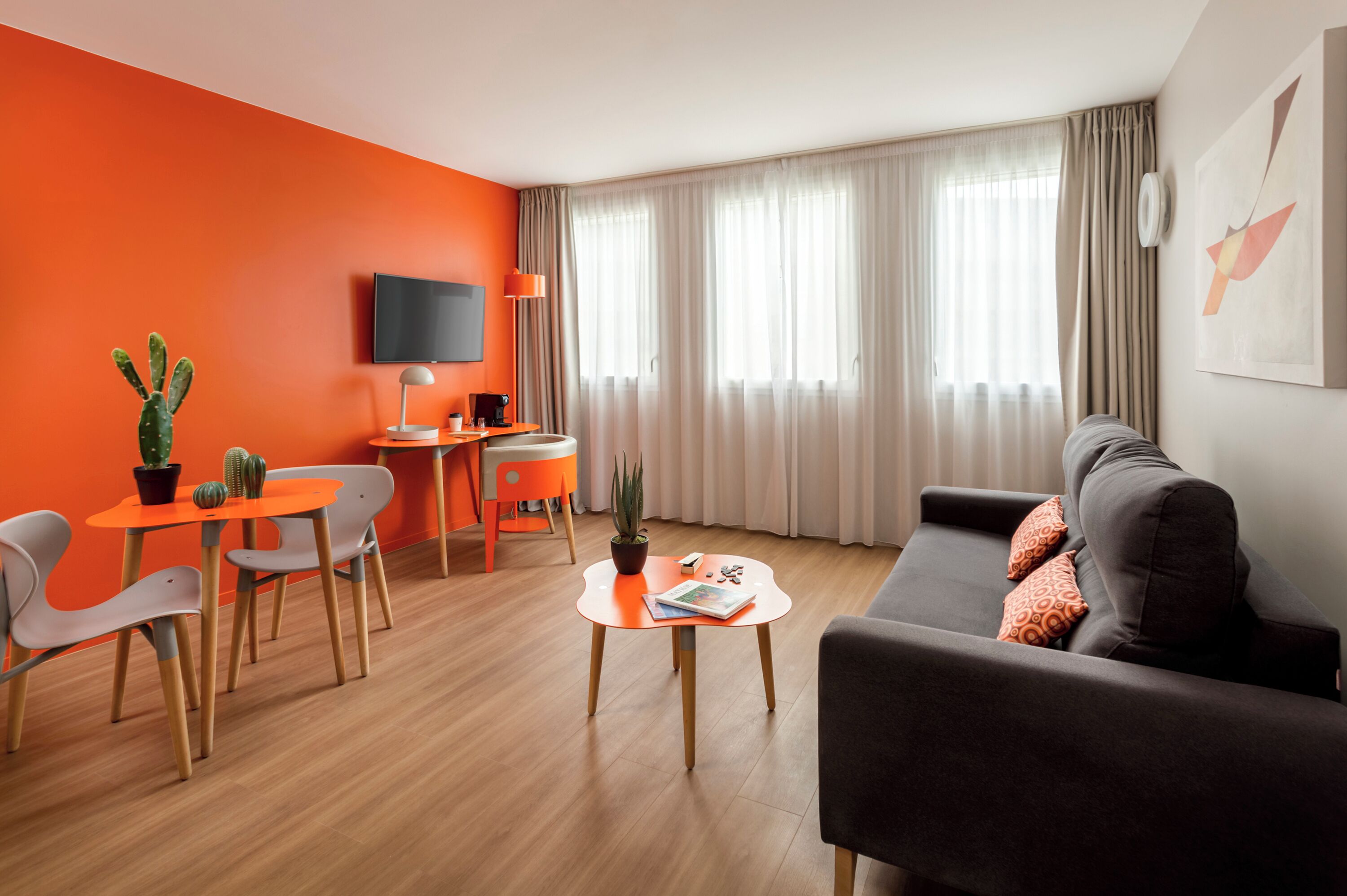 Un salon moderne avec un canapé, une table à manger et des chaises. Un téléviseur est fixé au mur, il y a une machine à café et un cactus sur la table.