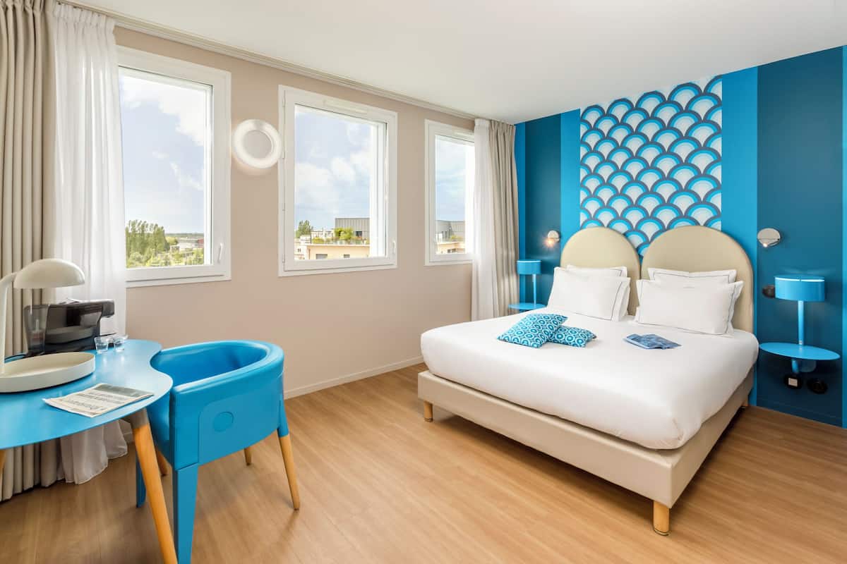 A bedroom with a bed, a desk, a chair, and a view of the outside.