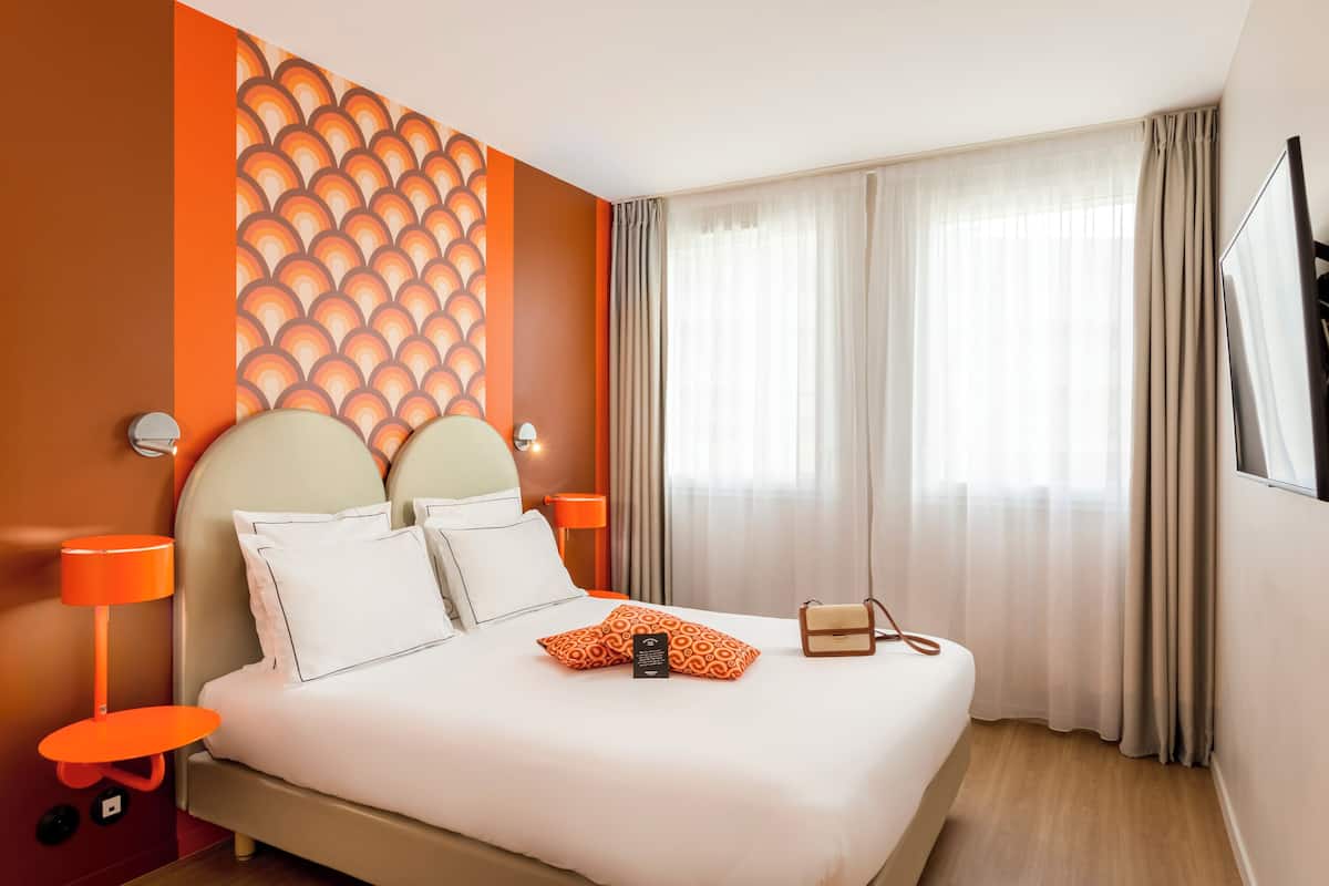 A hotel room with a large bed, two bedside tables, a wall-mounted TV, and a window with curtains.
