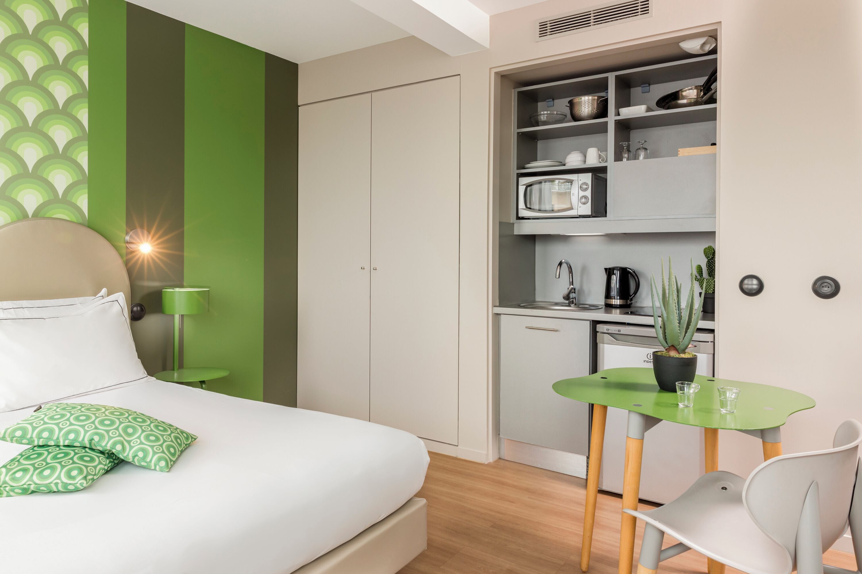 A compact bedroom with a bed, green patterned pillows, a green wall, a small table with a plant, and a kitchenette with a microwave and cabinets.
