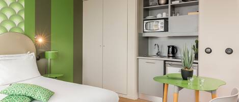 A compact bedroom with a bed, green patterned pillows, a green wall, a small table with a plant, and a kitchenette with a microwave and cabinets.