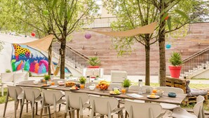 Un área de comedor al aire libre con terraza de madera, sillas blancas y una mesa servida con frutas, verduras y bebidas.