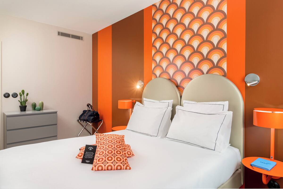 A hotel room with a bed, two bedside tables, a nightstand, a lamp, a suitcase, and a potted plant.