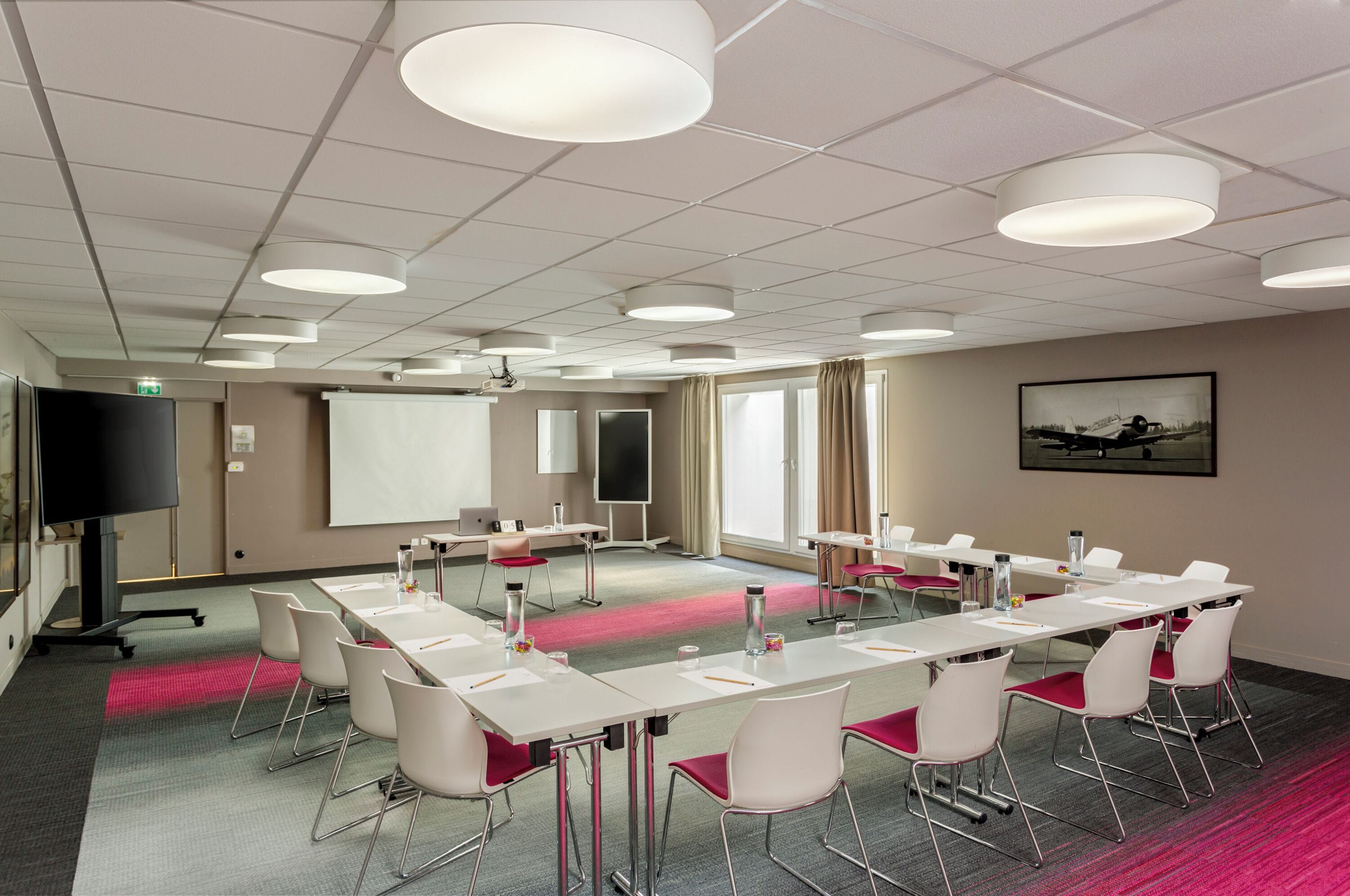 A conference room with a long table, white chairs, a projector screen, and a large window with curtains.