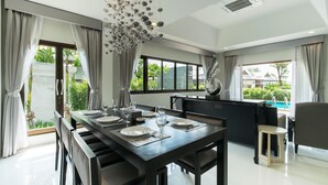 Un espace de repas moderne avec une table en bois foncé disposée pour quatre personnes, de grandes fenêtres offrant une vue sur la verdure et une piscine, et un lustre original agrémenté d’éléments sphériques.