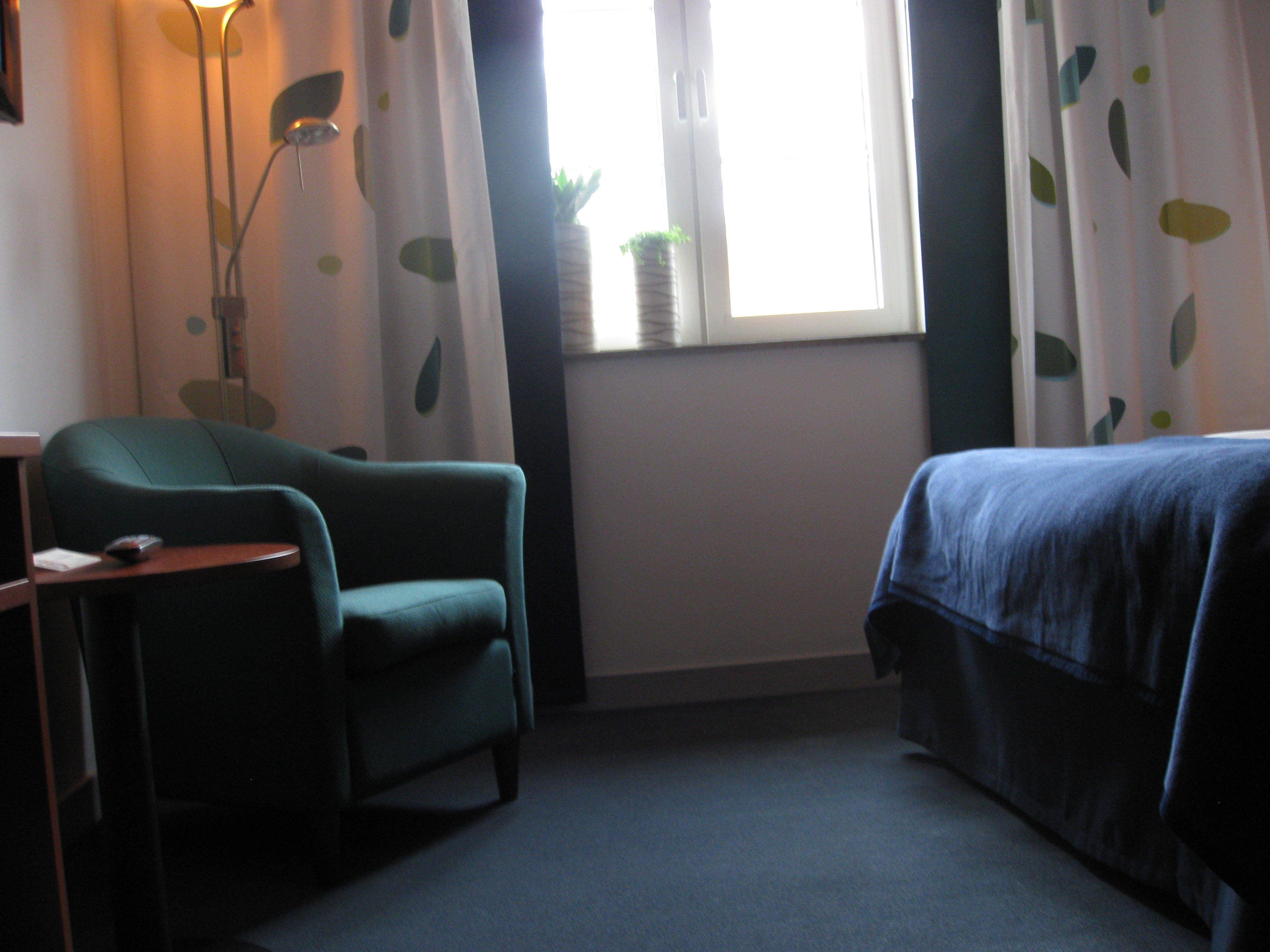 A room with a green armchair, a bed with a blue cover, a bedside table, a lamp, and a window with curtains.