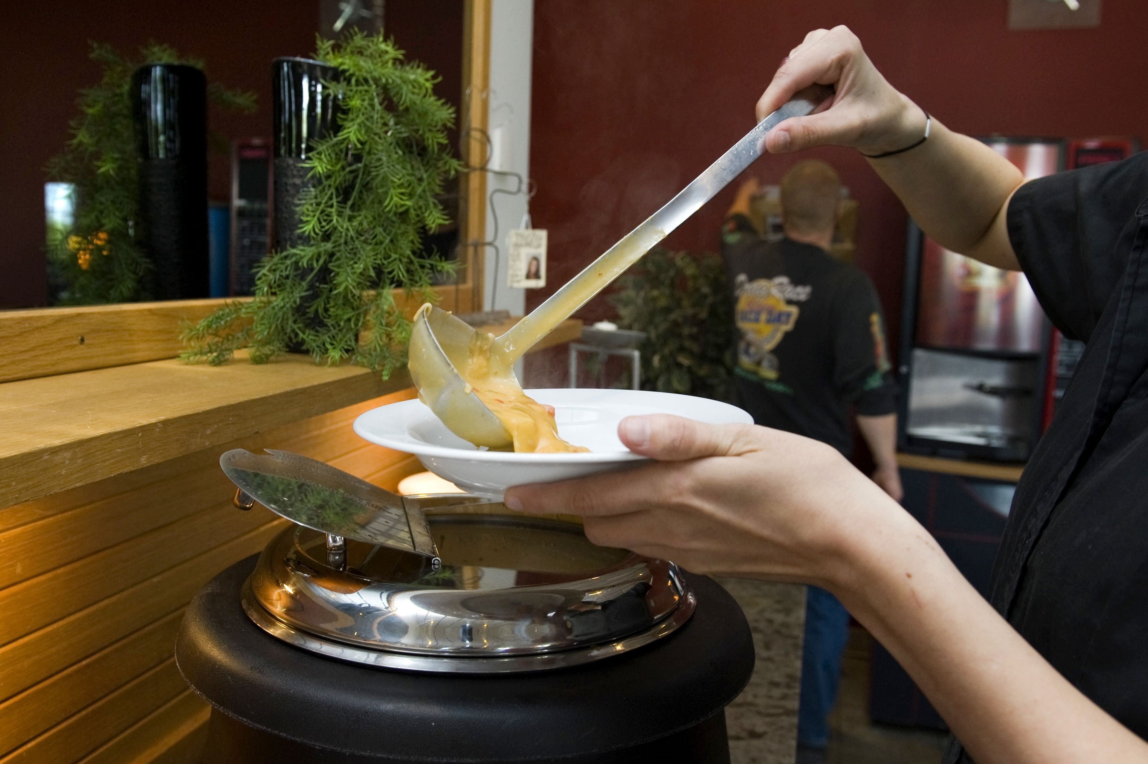 Una persona sosteniendo un plato con una cuchara, de pie cerca de un mostrador con una planta y un electrodoméstico metálico.