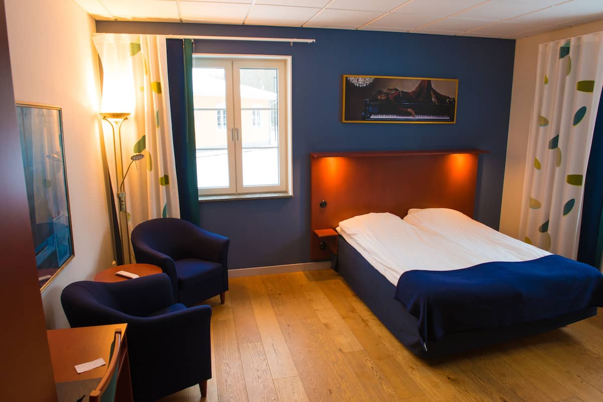 A hotel room with a bed, two armchairs, a desk, a lamp, and a painting on the wall.