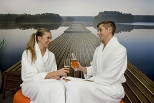 Duas pessoas em roupões brancos sentadas em um píer de madeira, cada uma com uma cerveja, apreciando um lago calmo com um píer que se estende na água.