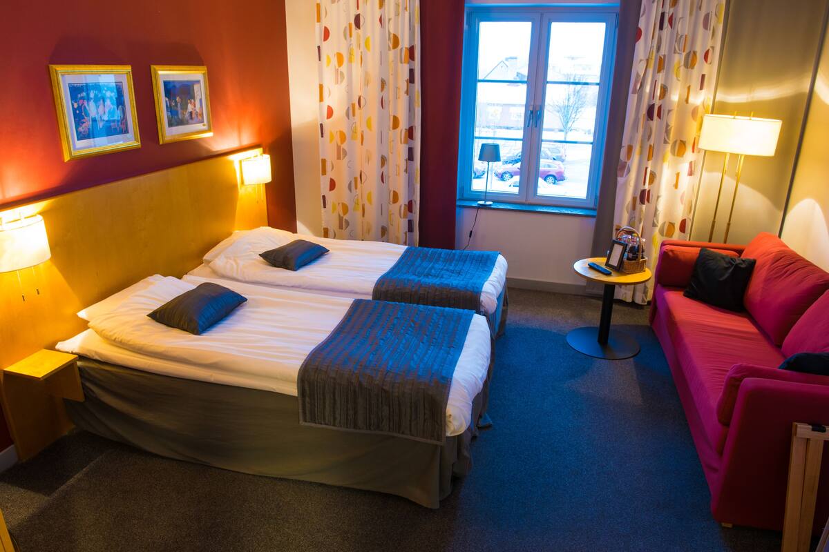 A hotel room with two beds, a red sofa, a small table, and a window with curtains.