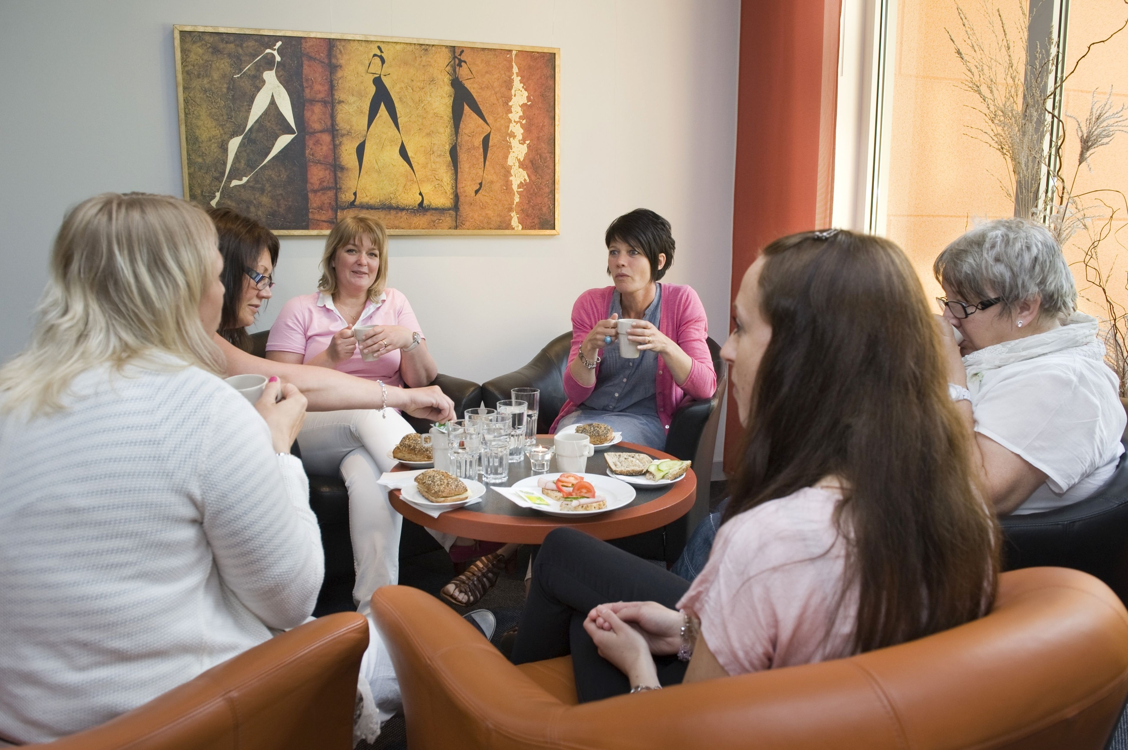 A group of people seated around a round table with food and drinks, in a room with a large abstract painting on the wall.