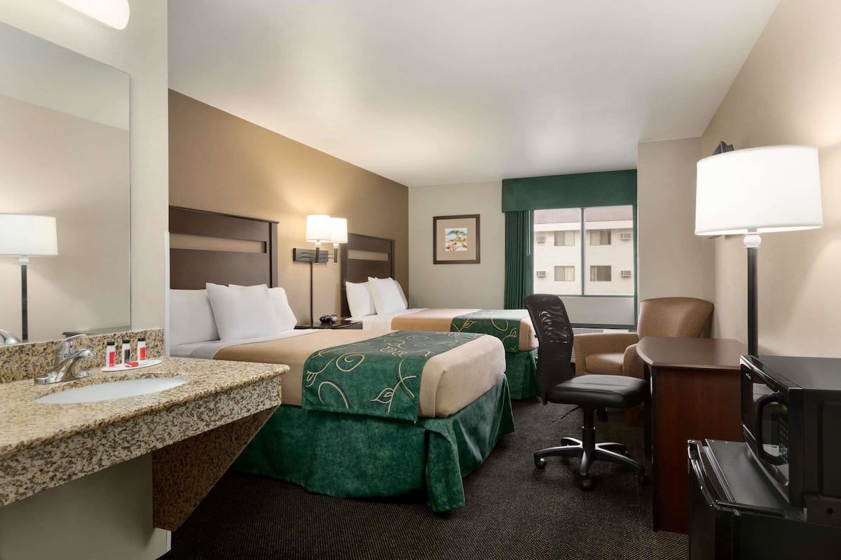 Une chambre d’hôtel avec deux lits, un bureau, une chaise, une lampe de bureau, une télévision et une fenêtre donnant sur des immeubles.