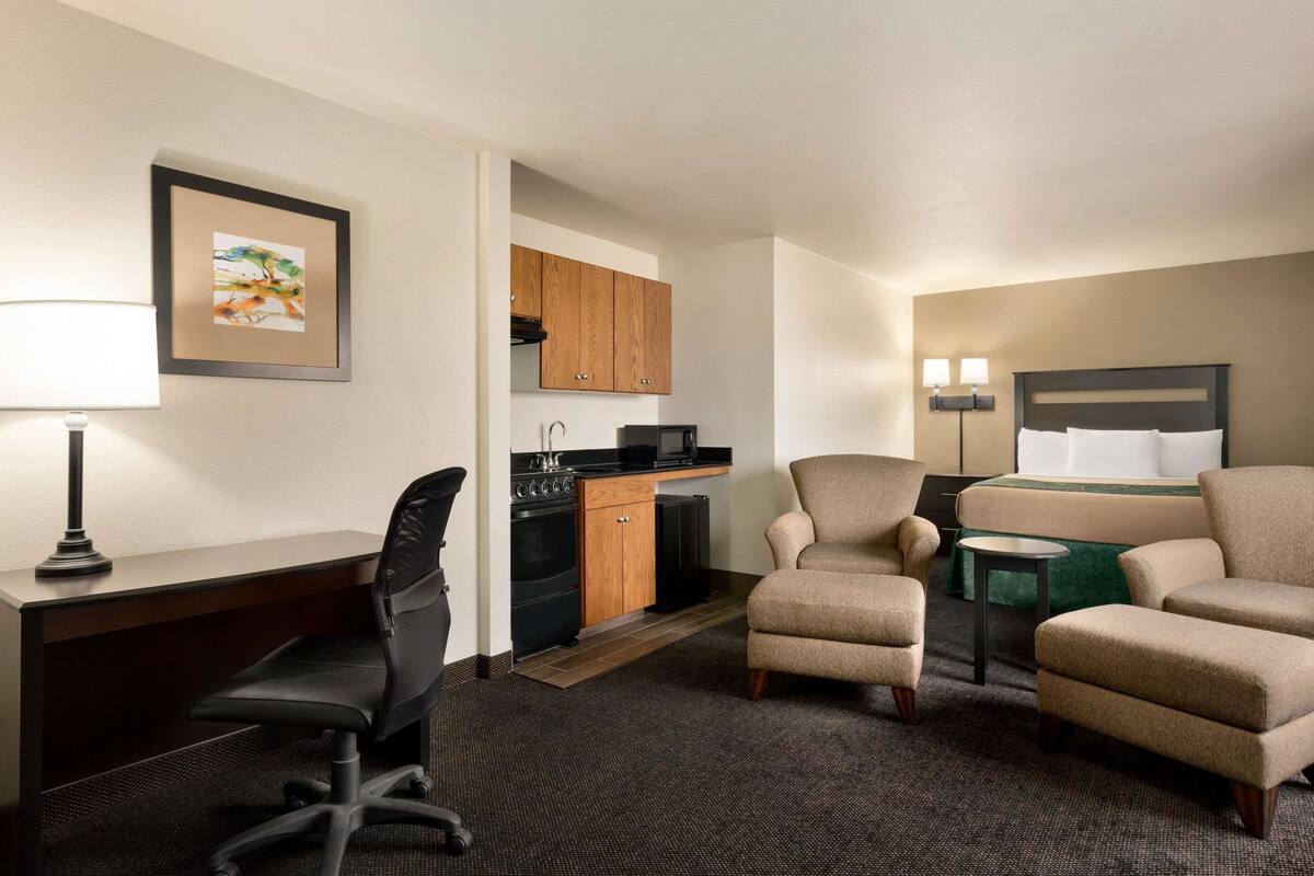 Une chambre d’hôtel équipée d’un bureau, d’un fauteuil de bureau, d’une kitchenette, de deux fauteuils, d’un lit et d’une petite table.