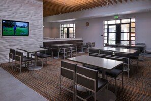 A modern dining area with multiple tables and chairs, a large screen displaying a golf game, and large windows providing natural light. - Residence Inn Palo Alto Menlo Park (Menlo Park)