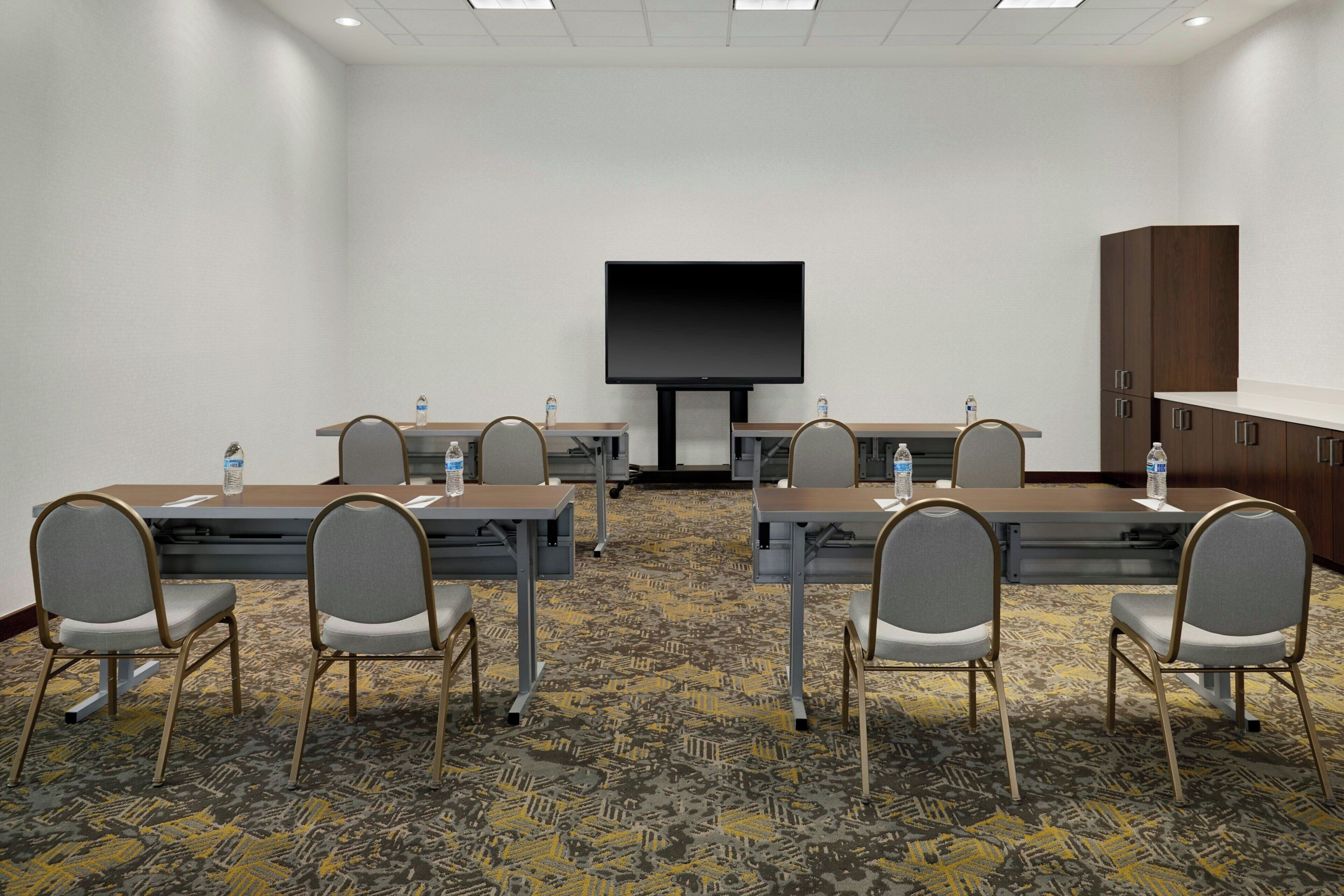 A conference room with six tables, each with two chairs, a large flat-screen TV, and water bottles on the tables.